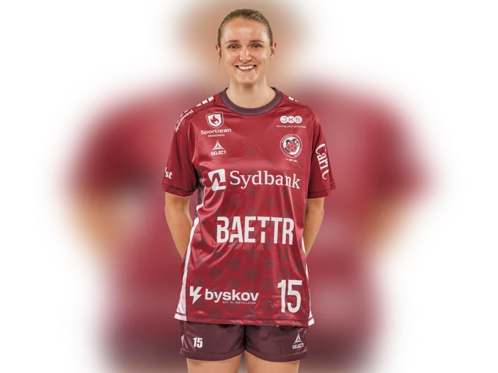En kvinde i en rødbrun sportsdragt med nummer 15 står smilende med hænderne bag ryggen foran en sløret hvid baggrund.